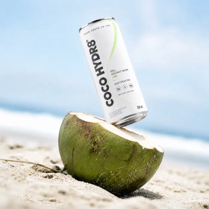 A can of COCO HYDR8 Lime balanced on a split green coconut on a sunny beach