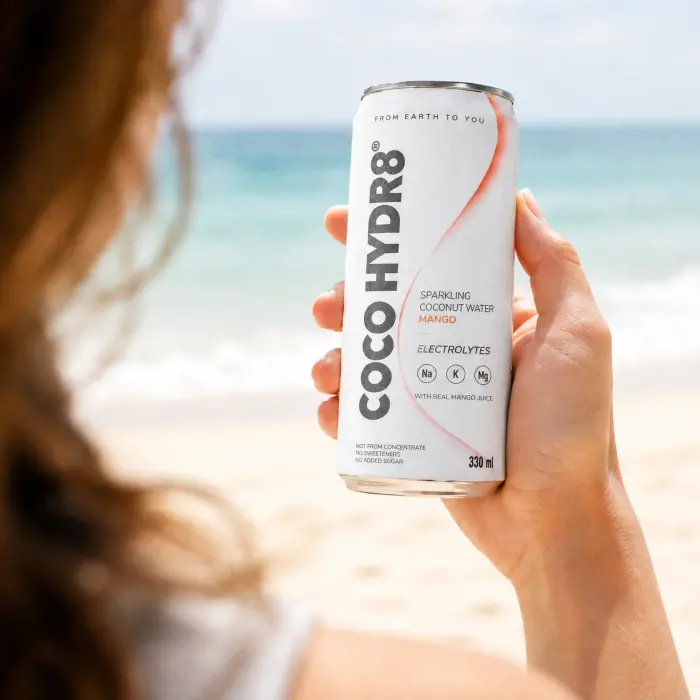 A hand holding a can of COCO HYDR8 Mango on a sunny beach