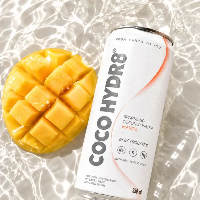 A can of COCO HYDR8 Mango beside a scored mango in sunlit water