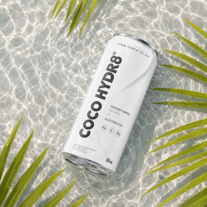 A can of COCO HYDR8 Natural lying in clear, sunlit water between palm fronds