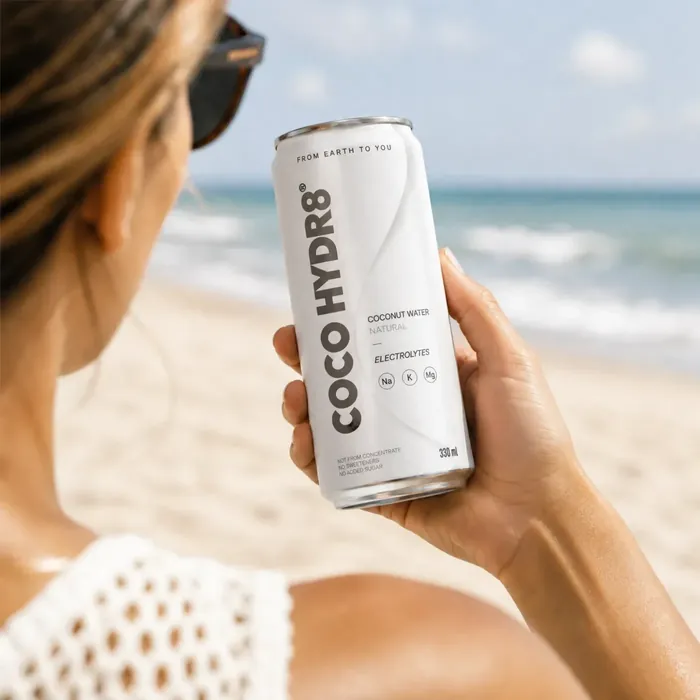 A woman in sunglasses holding a can of COCO HYDR8 Natural at the water's edge